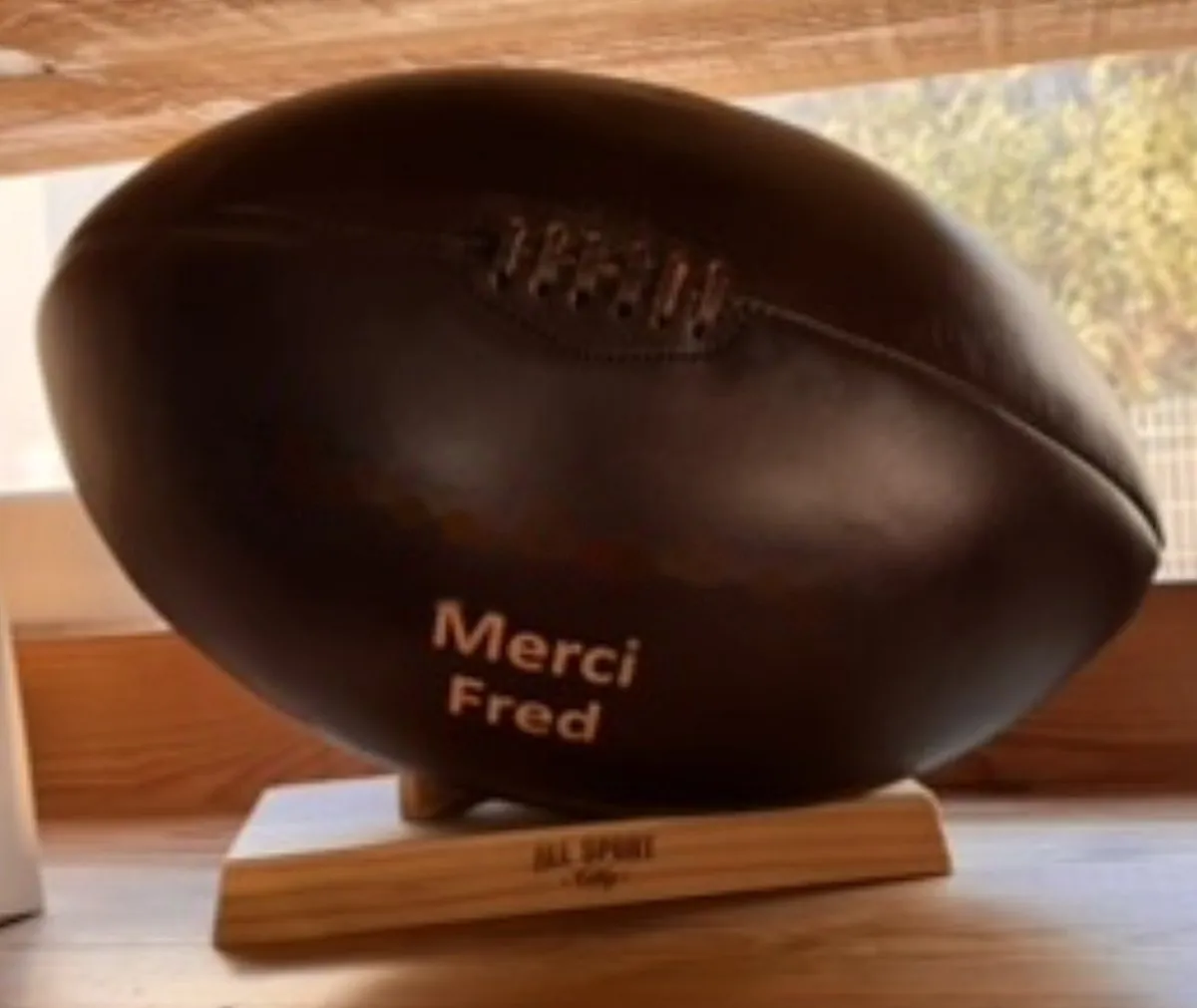 Un ballon de rugby en cuir sombre posé sur son socle en bois, gravé « Merci Fred »