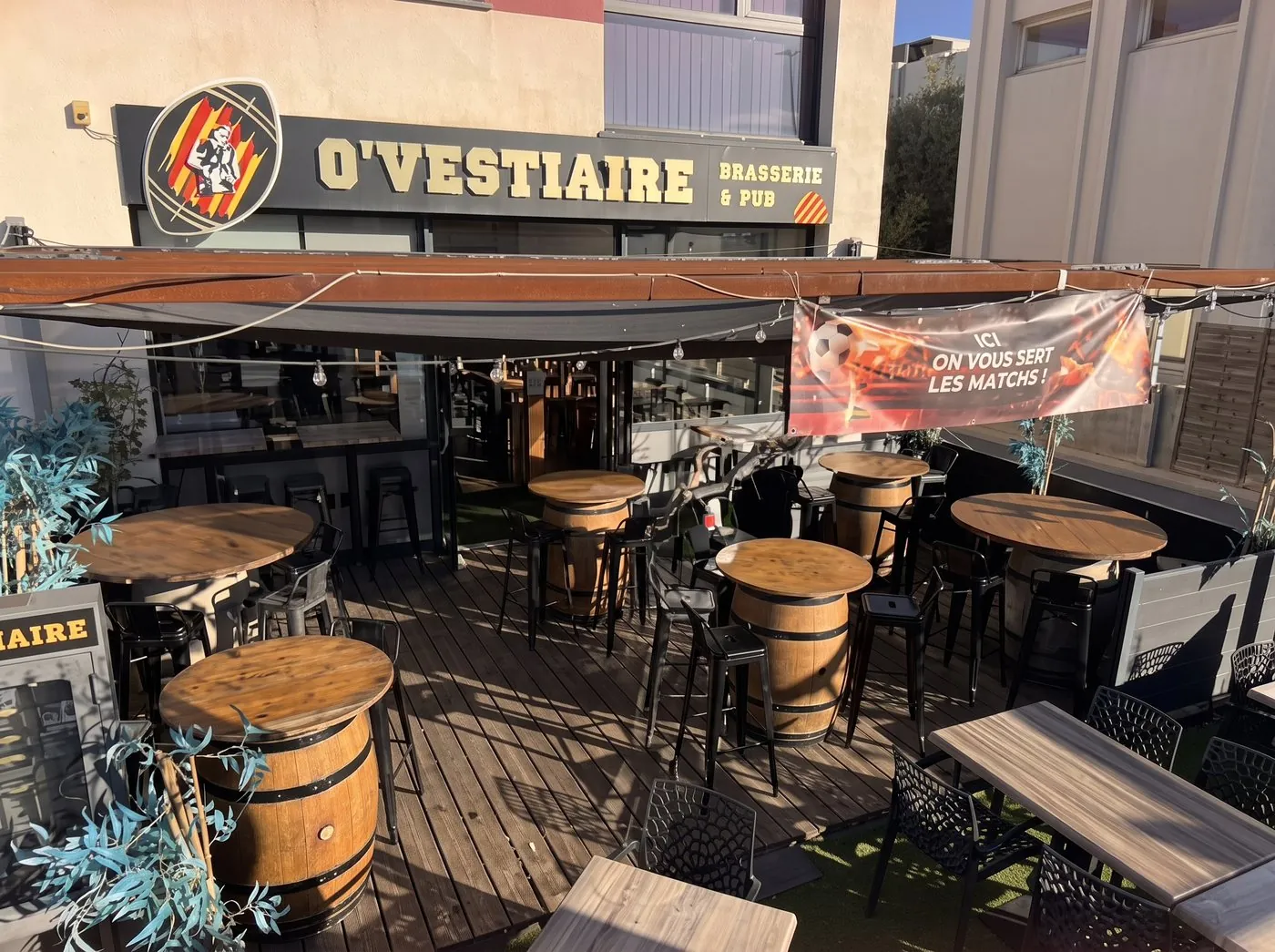 La terrasse d’O’Vestiaire sous l’auvent : tables-tonneaux, tabourets et plancher en bois, au soleil